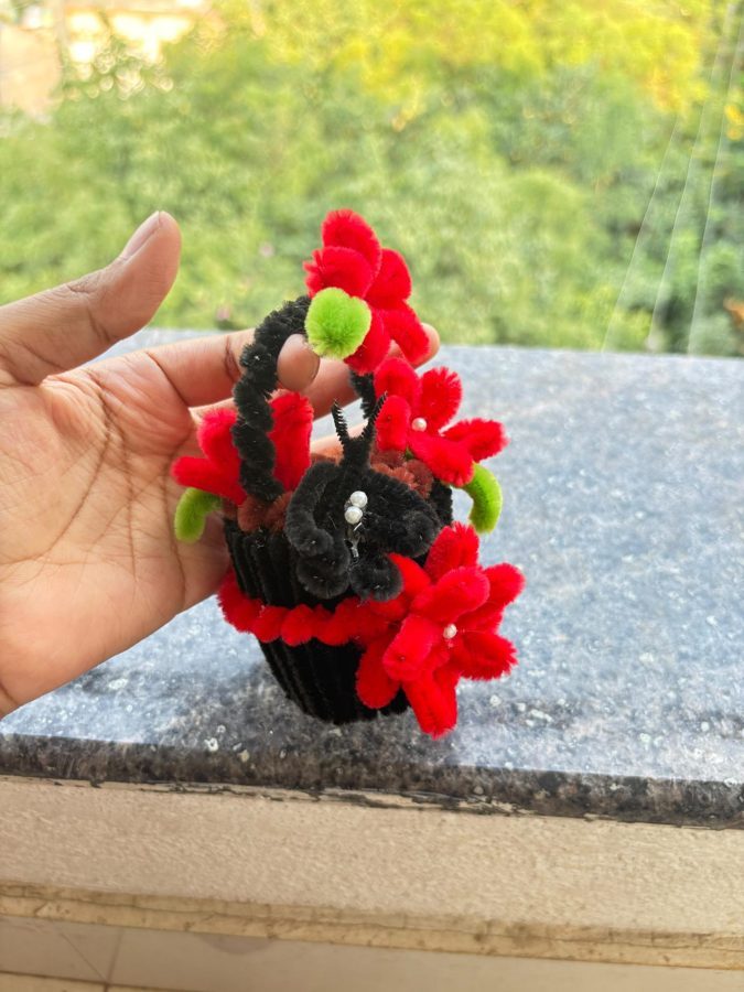 Ruby Butterfly Basket. Handcrafted mini flower basket made from soft chenille wire, with red blossoms, pearl centers, and green leaves. Features a black handle, a little black butterfly, and a red-trimmed knitted base…
